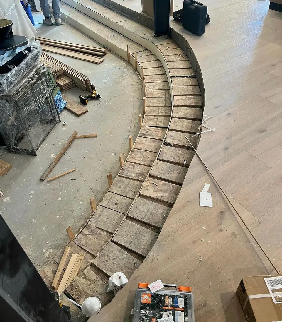 Curved plank run being built around a stair base on an open subfloor